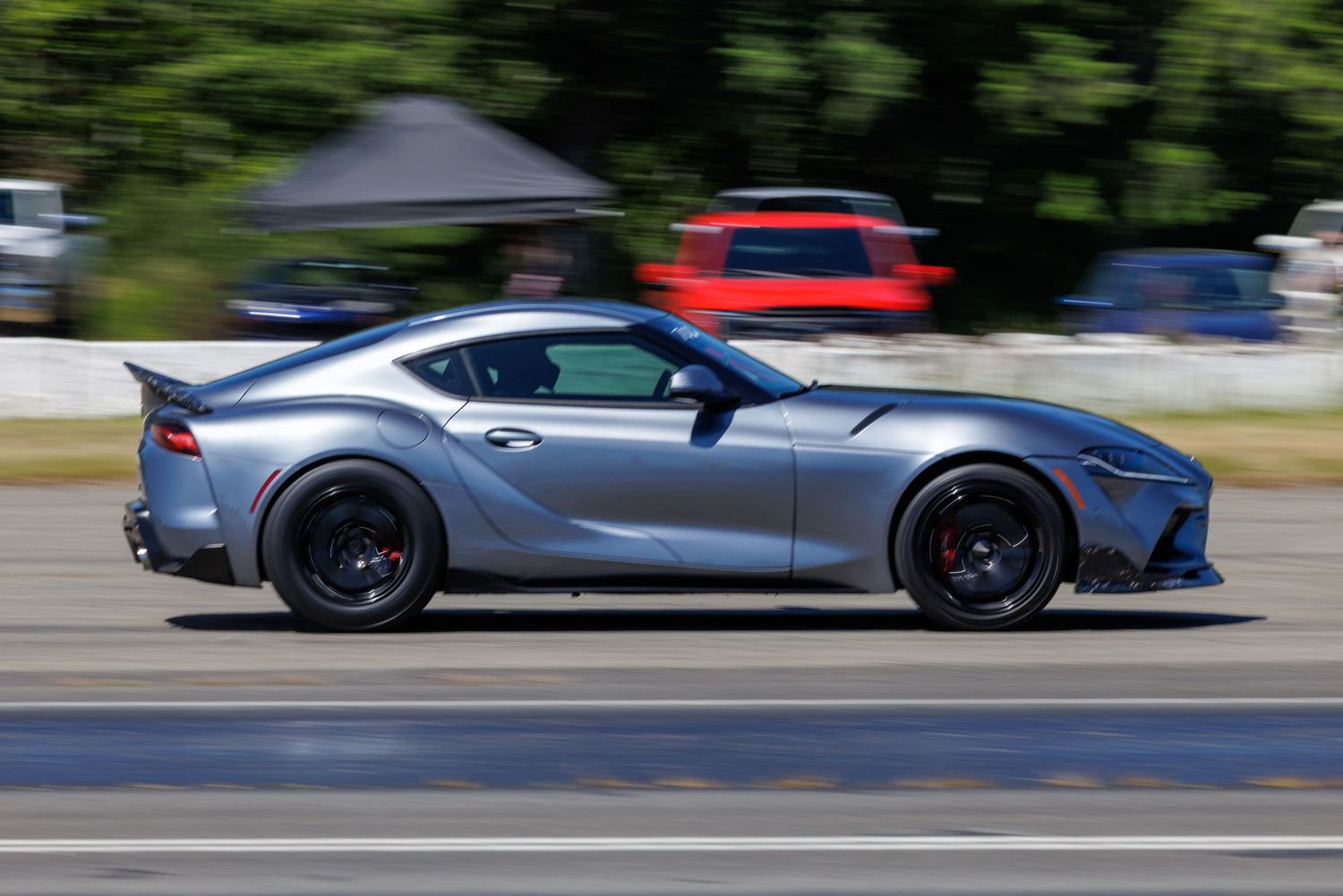A silver Toyota Supra sports car speeding on a track with motion-blurred background trees and a red car visible.
