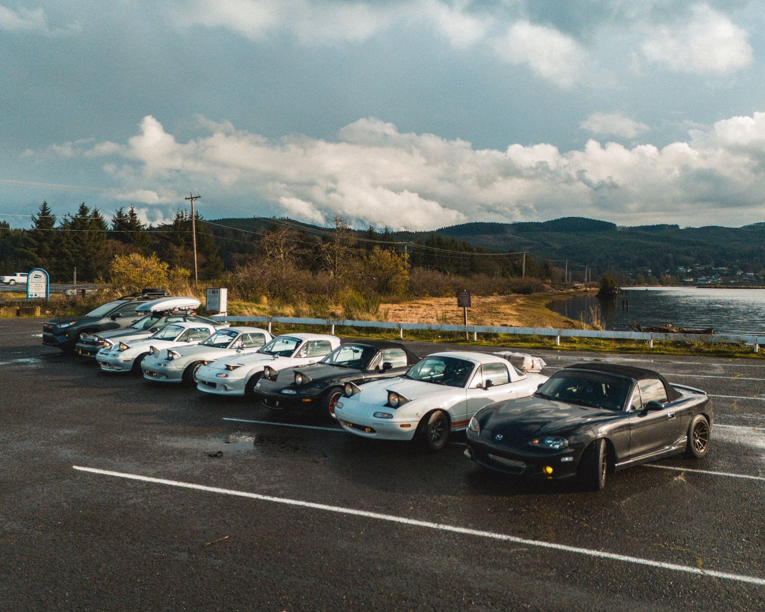 Row of vintage cars parked in a lot with mountains and cloudy sky in the background.