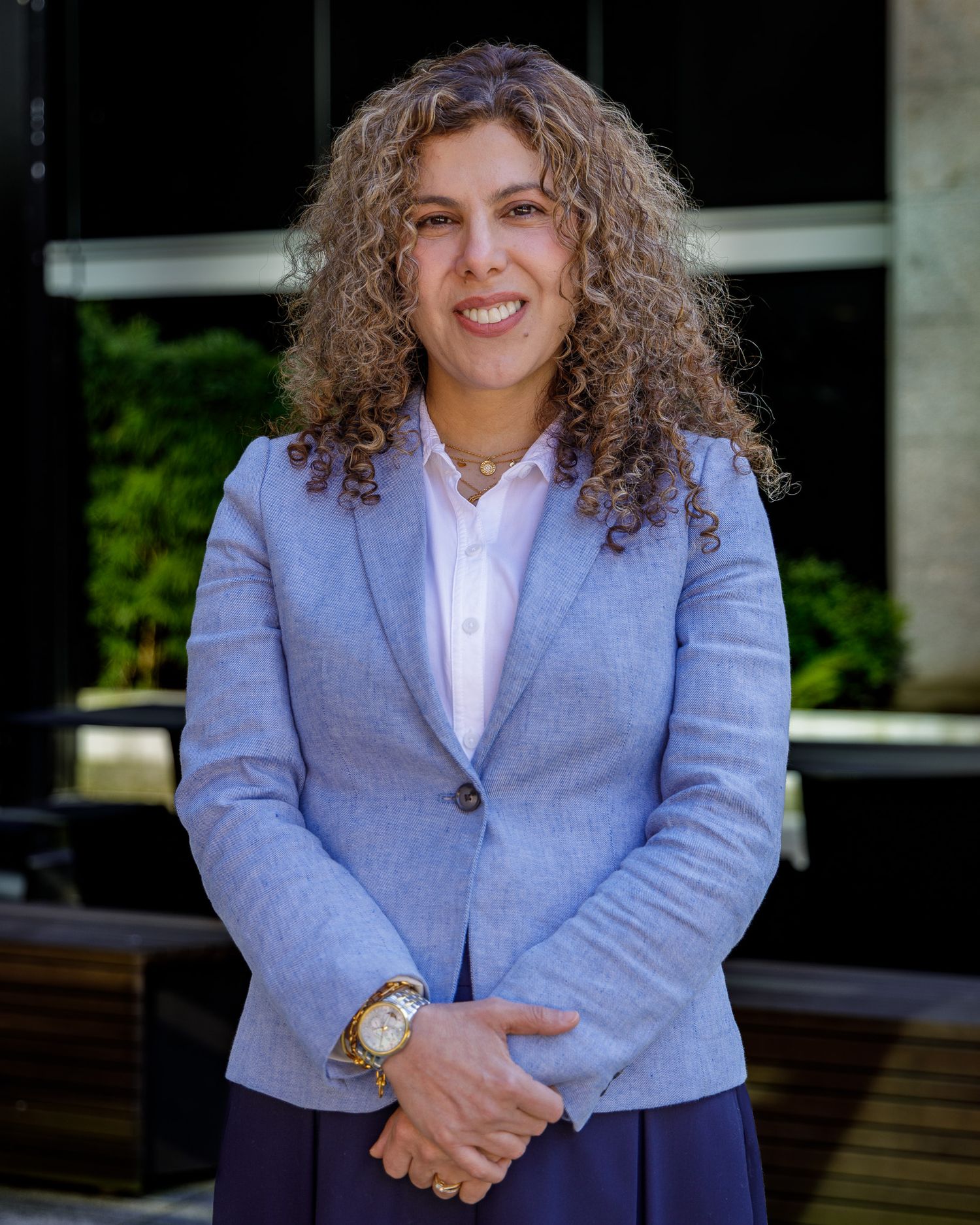 Professional portrait of a person in light blue blazer with curly hair against office background.