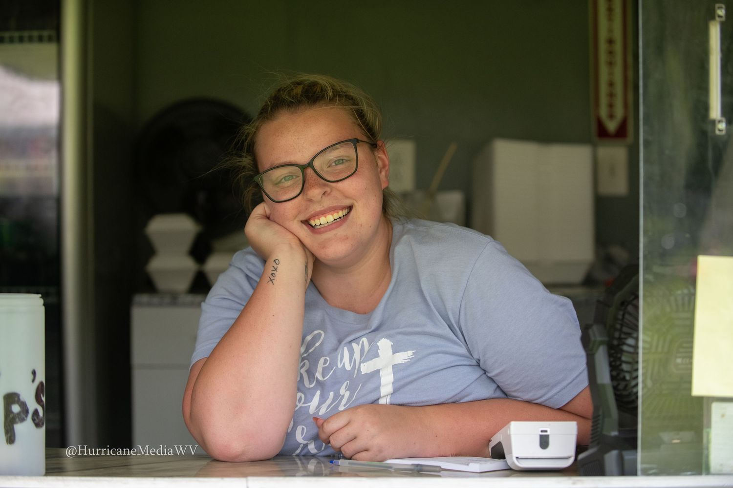 A person wearing glasses sits laughing at a table in front of a window.