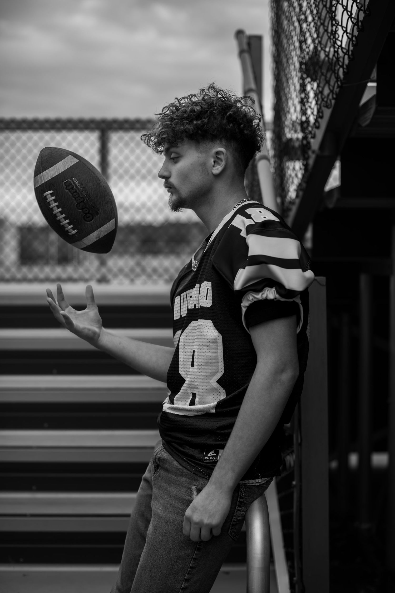 Black and white portrait of a football player in jersey number 8 tossing a football in the air near bleachers.