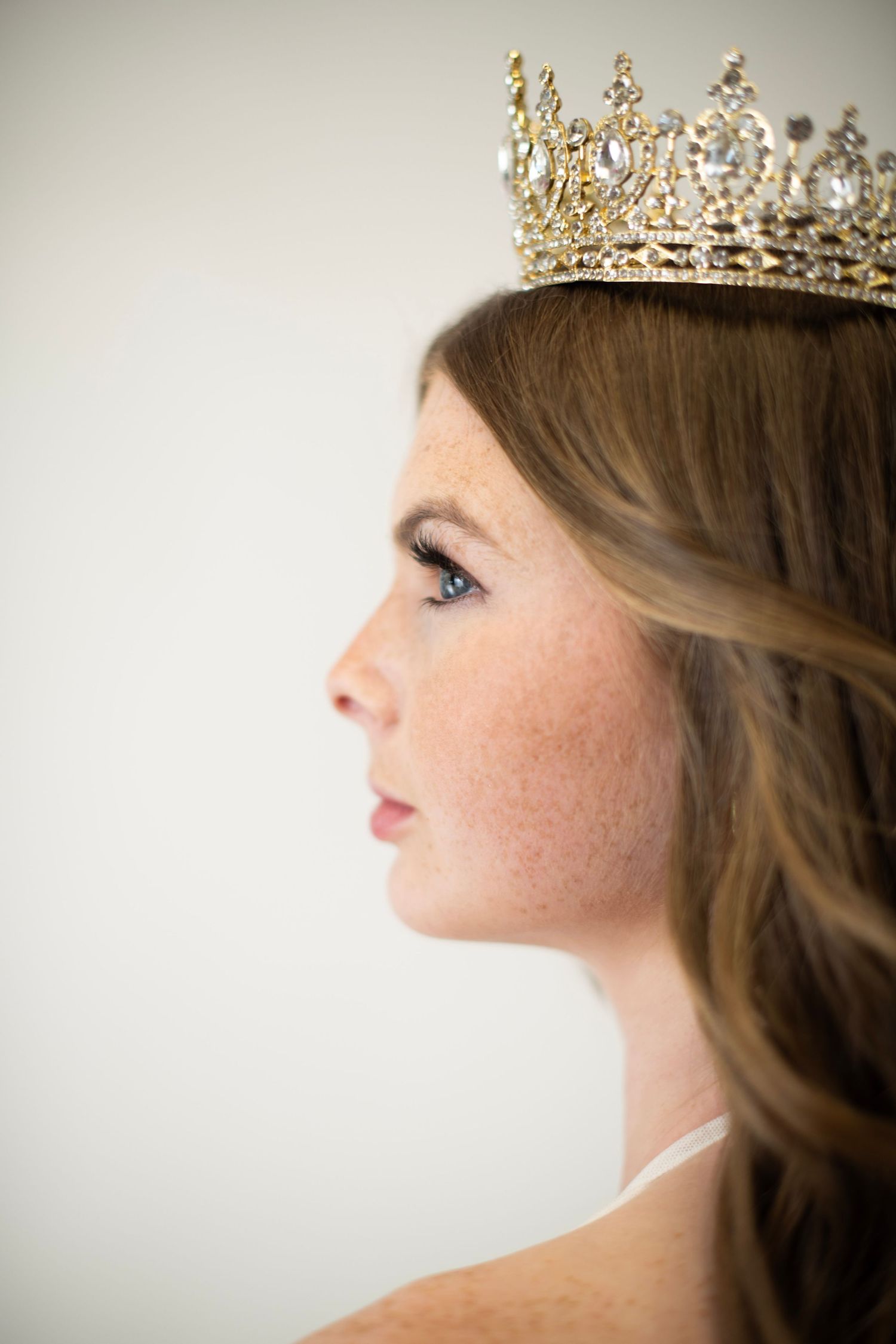Young woman wearing a gold crystal tiara crown, side profile view with long brown hair and freckles.