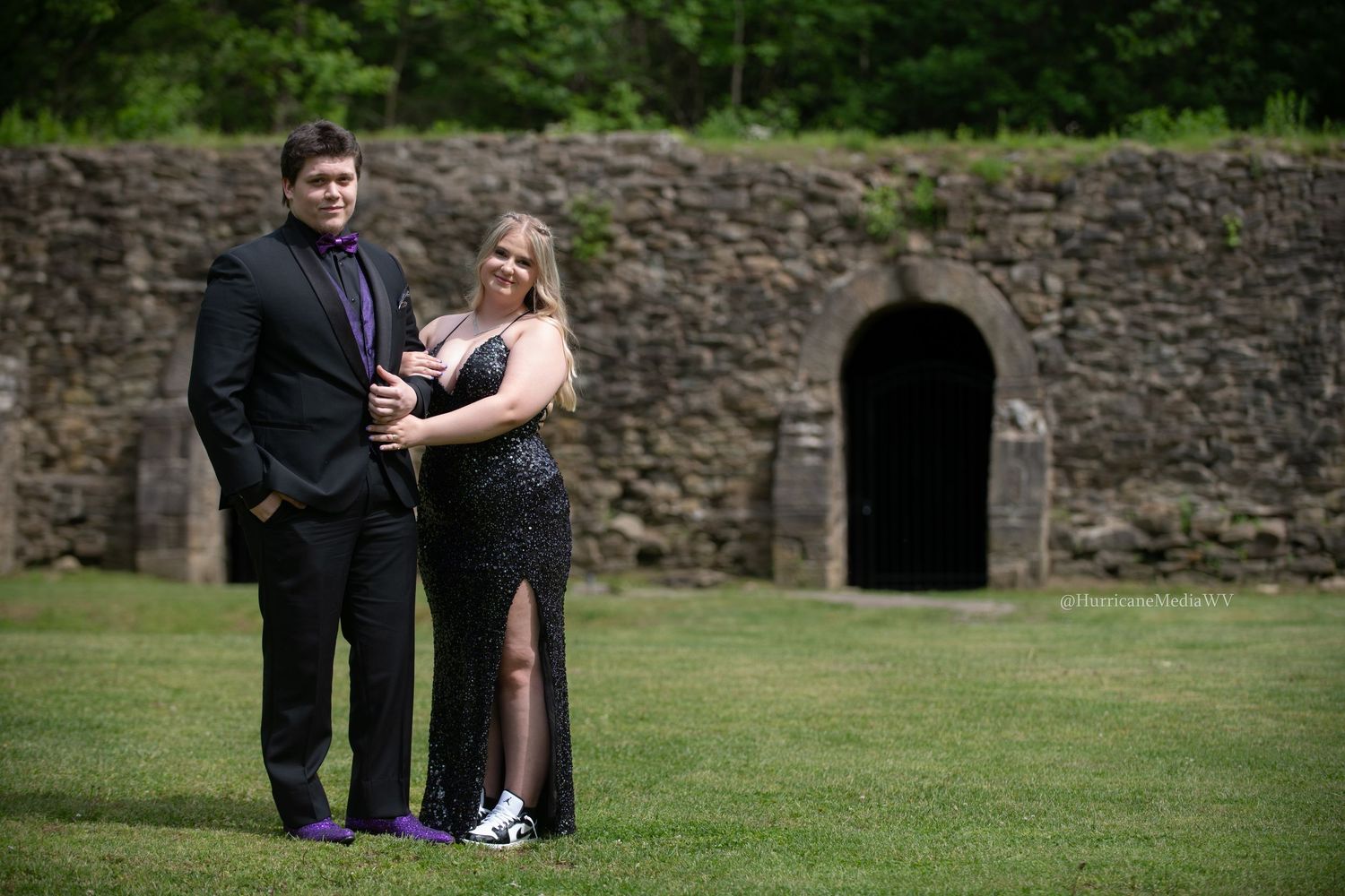 Couple in formal wear posing near stone wall with archway in garden setting.