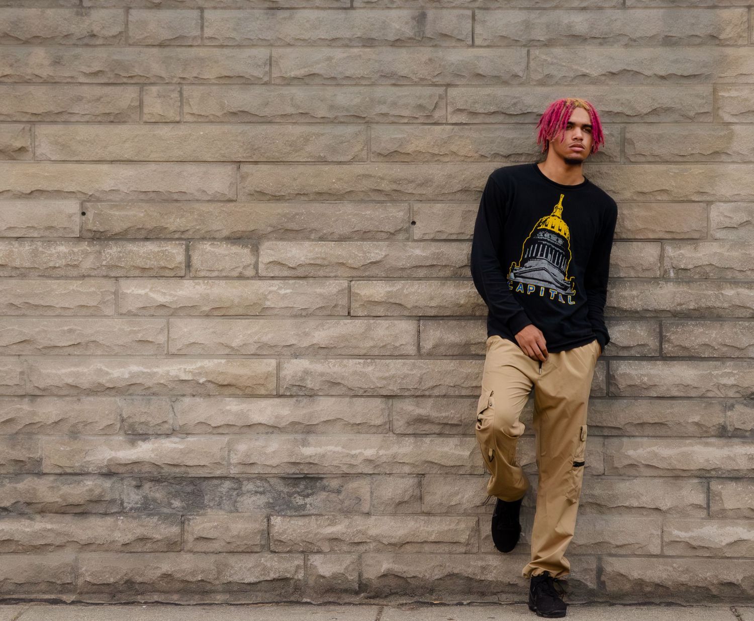 Person with bright pink hair leaning against textured stone wall wearing graphic t-shirt.