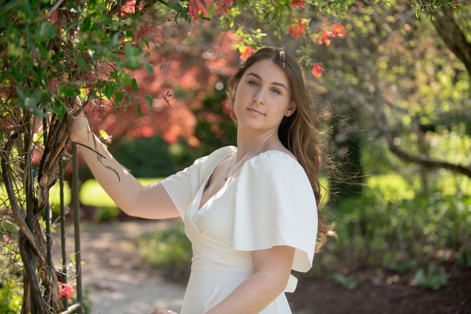 Person in white dress poses elegantly near flowering tree in garden setting.