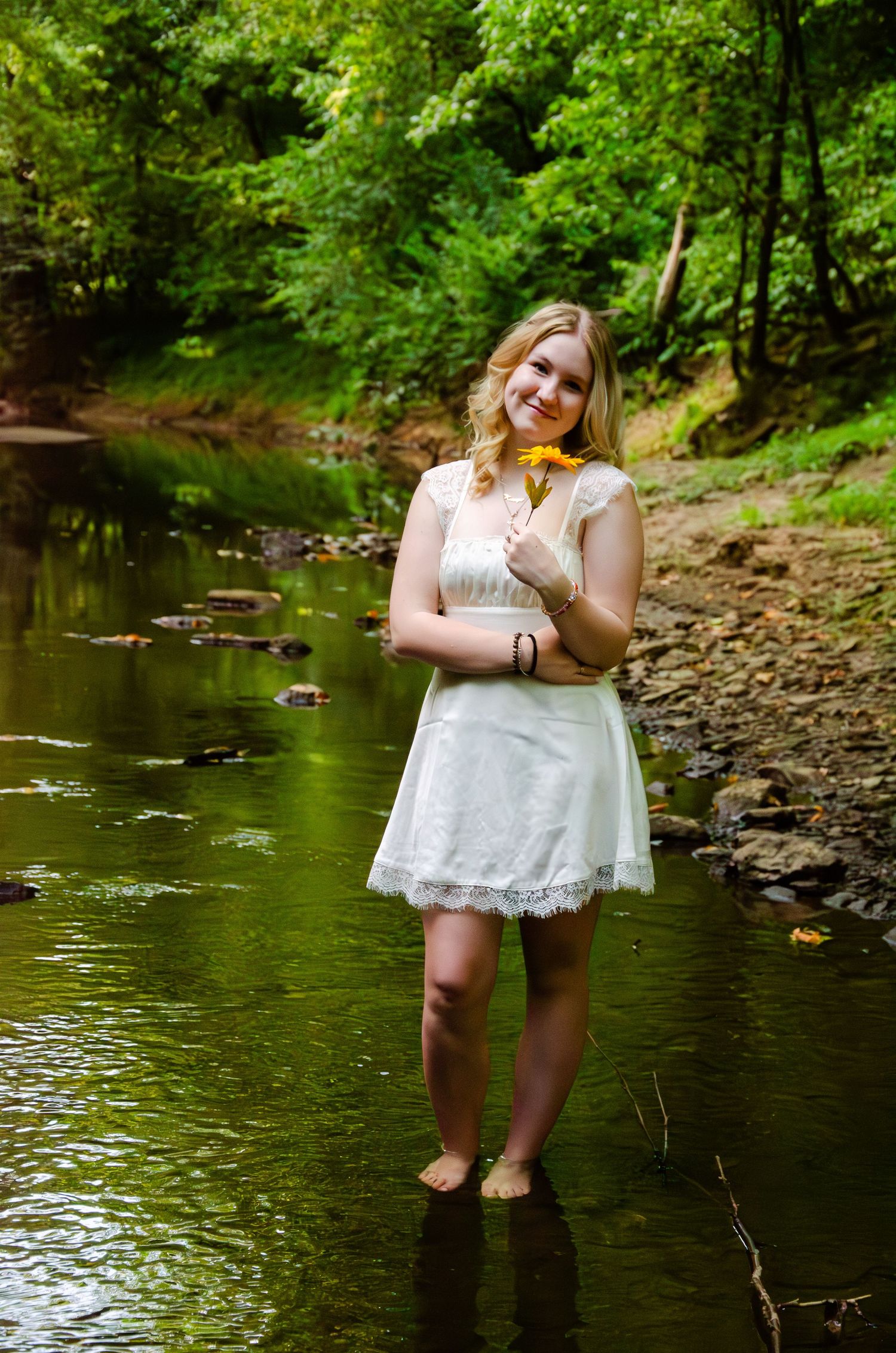 Someone in a white lace dress stands barefoot in a tranquil creek surrounded by lush green forest scenery.