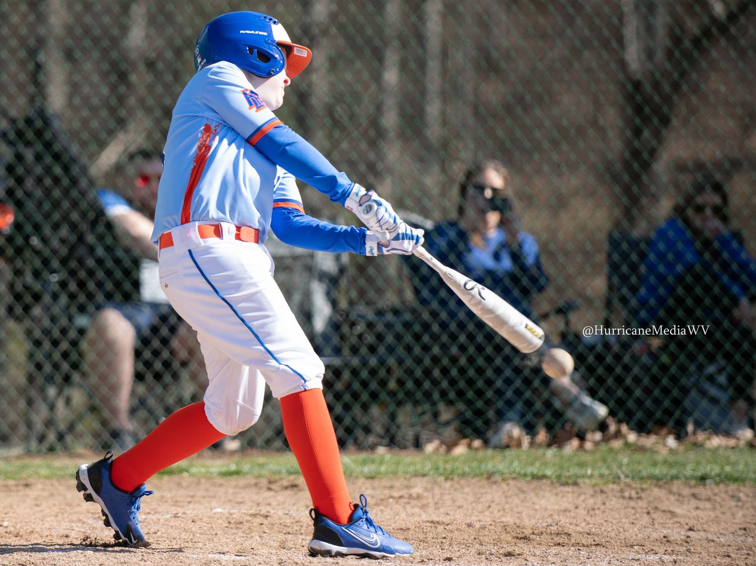 Baseball batter in patriotic uniform swinging bat during game through chain link fence.