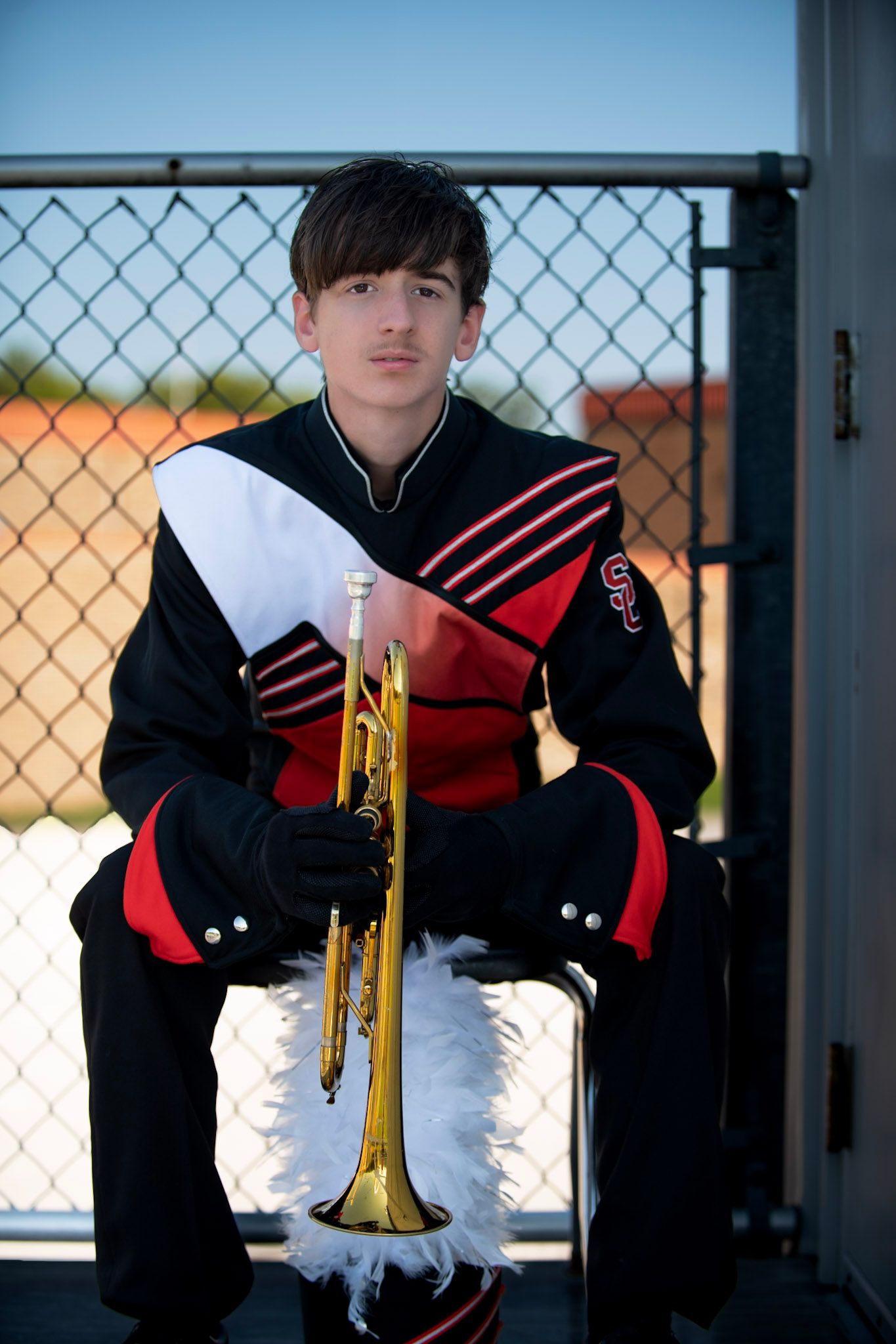 Band member in black and red uniform poses with trumpet against chain-link fence background.
