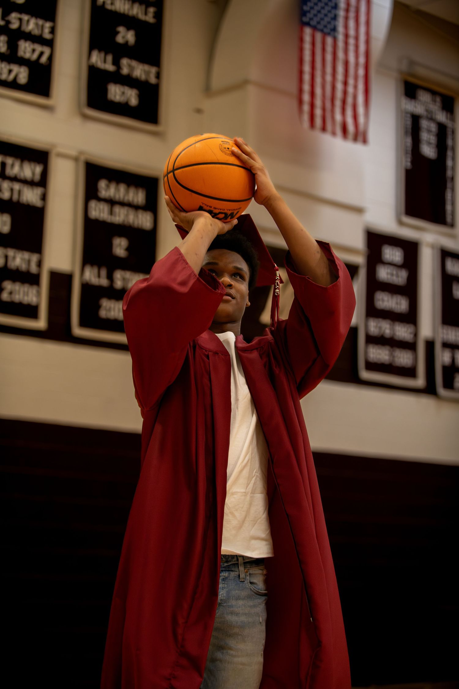 Basketball player in maroon jacket practices shooting in gym with American flag on wall.