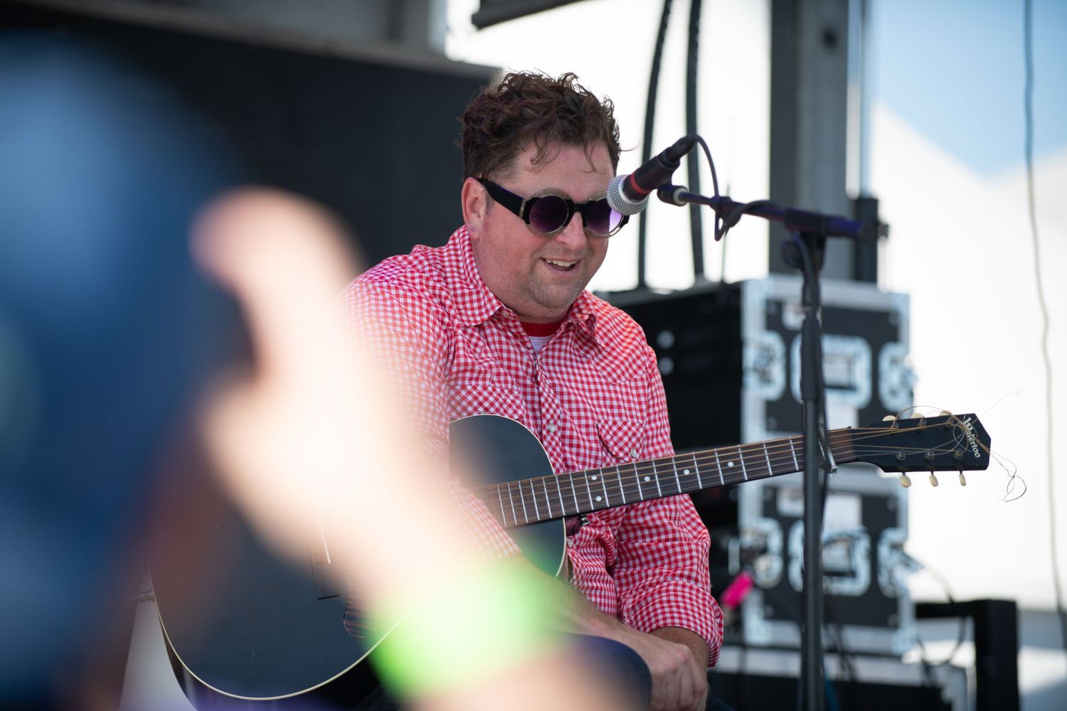 Musician in red checkered shirt plays acoustic guitar on outdoor stage during daytime performance.