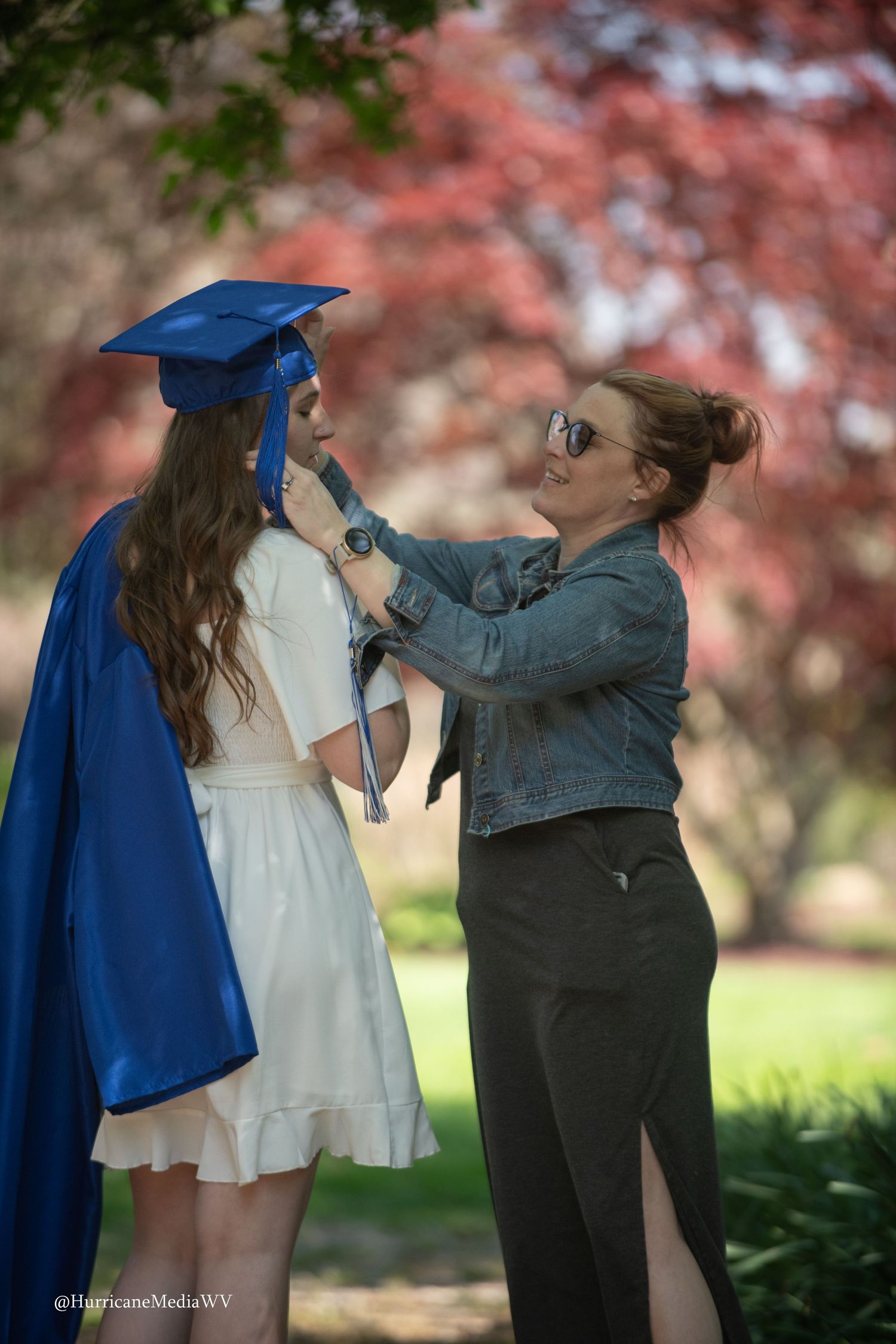 A woman in a blue graduation gown and someone in casual clothes interacting joyfully outdoors with pink blossoms in background.