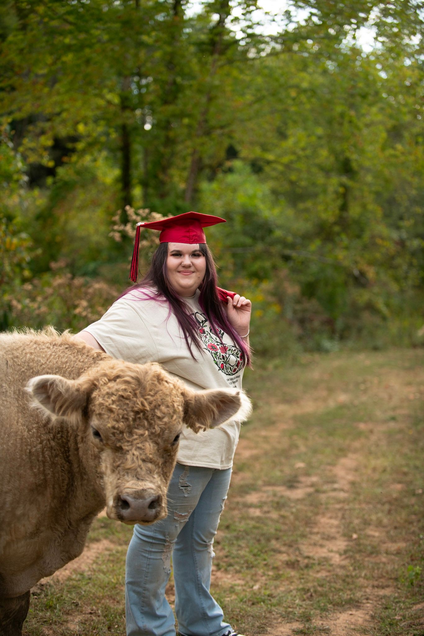Graduate in red cap poses with brown cow in outdoor country setting.