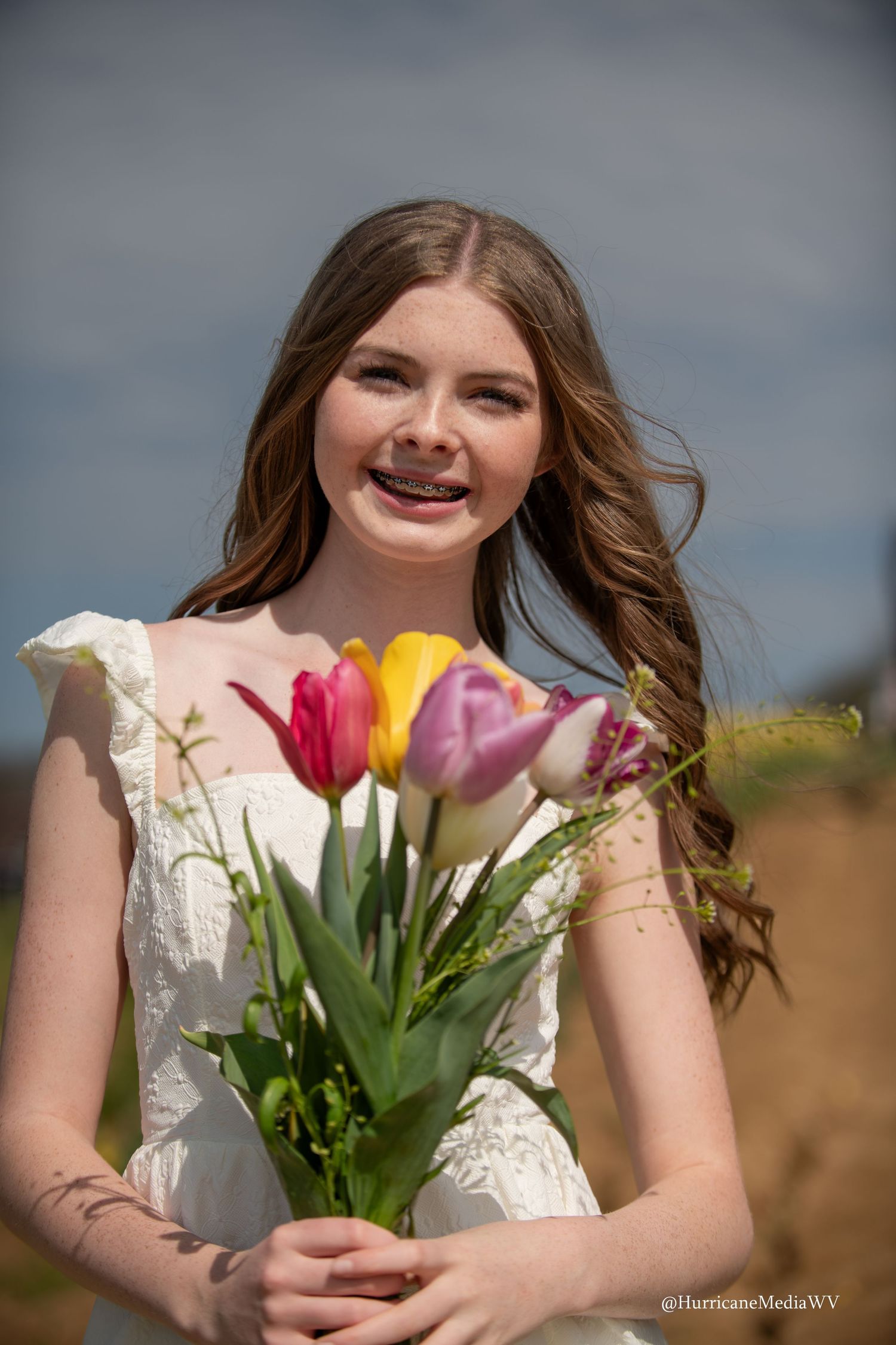 Person in white dress holds colorful spring tulips against cloudy sky backdrop.