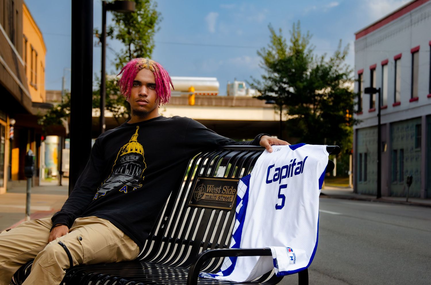 Person with pink hair holding basketball jersey while sitting on bench in urban setting.