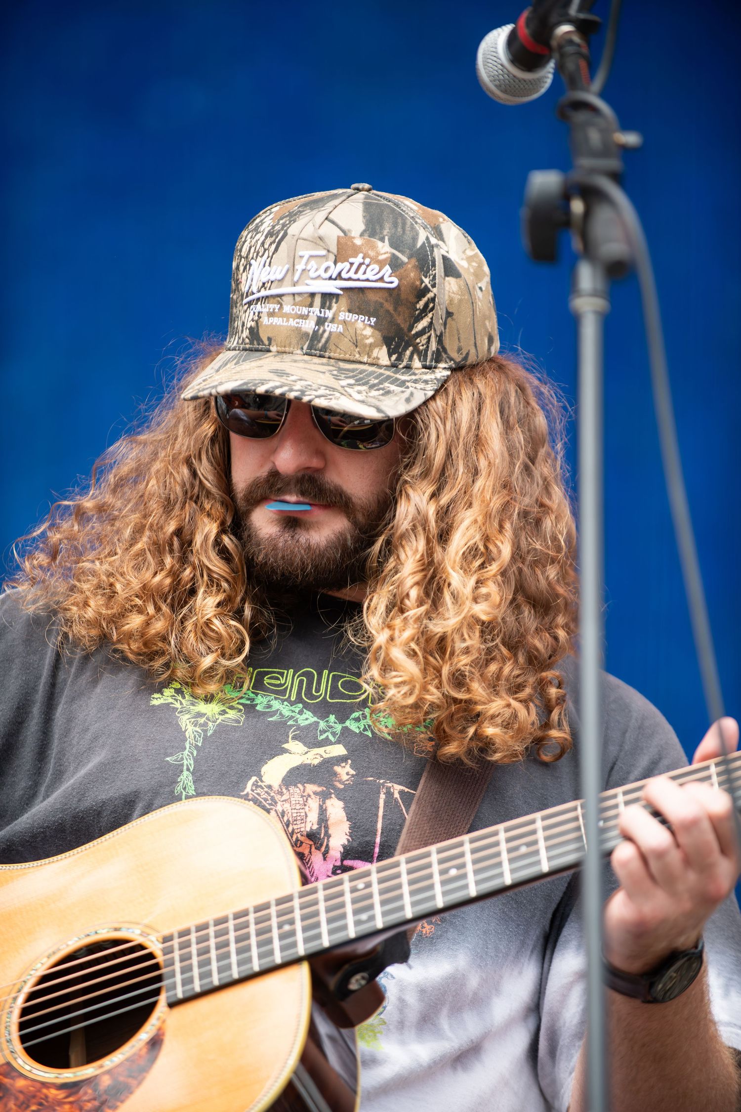Musician with long curly hair wearing camo hat and sunglasses plays acoustic guitar against blue sky background.