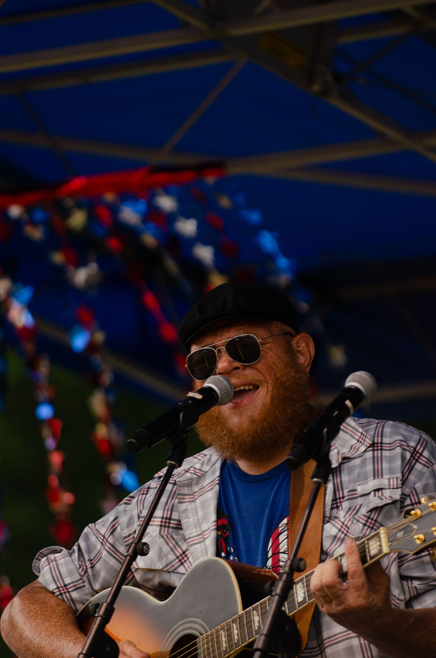 Musician performing with an acoustic guitar under a blue tent at an outdoor event.
