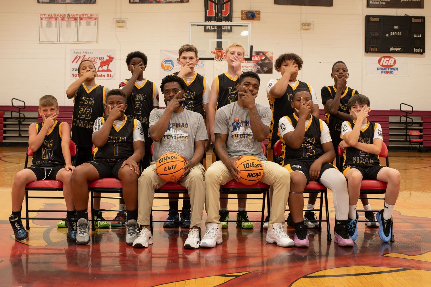 Basketball team members pose for a group photo sitting on chairs in a gym.