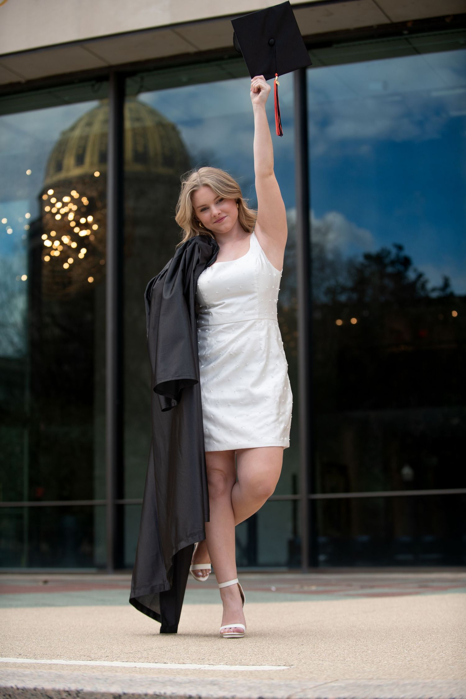 Graduate in white dress celebrating with graduation gown held high against glass building.