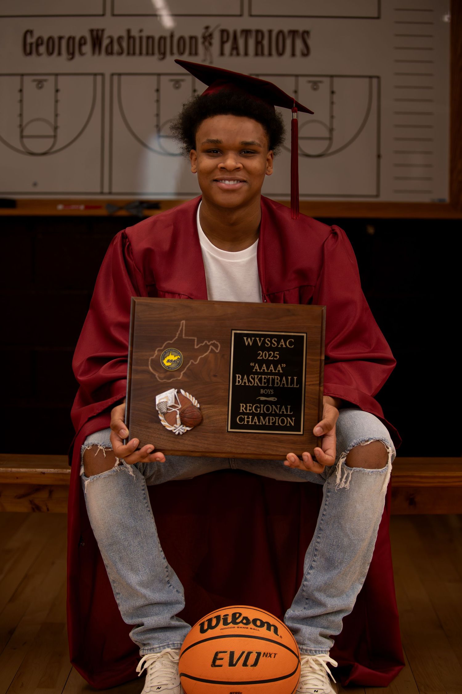 Graduate in red cap and gown displays basketball championship plaque in school gymnasium.