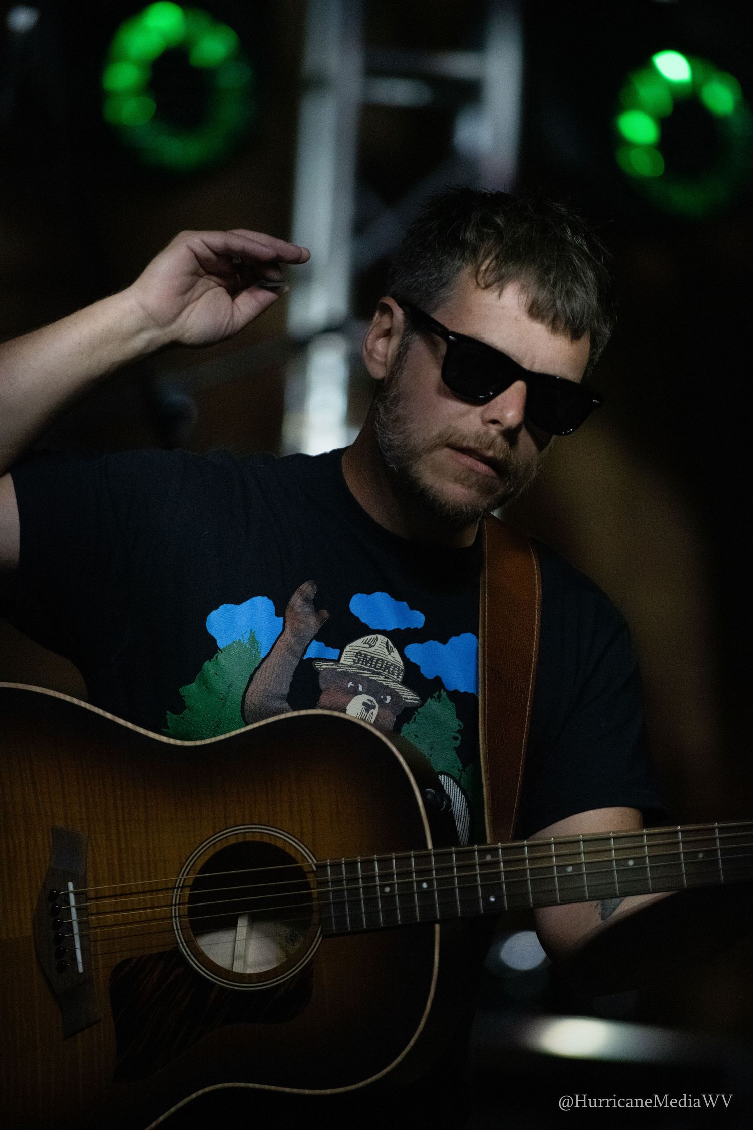 Musician plays acoustic guitar in dark setting with green lighting and soft bokeh effects in background.