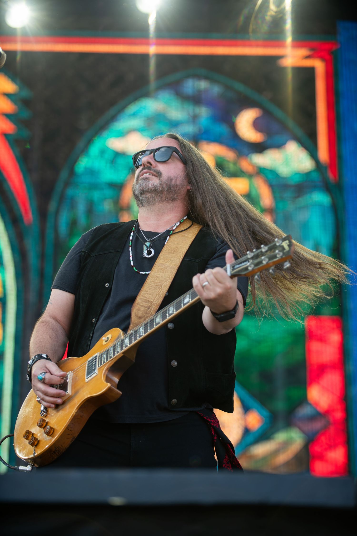 A guitarist with long hair performs energetically on stage with colorful blue and red lighting in the background.