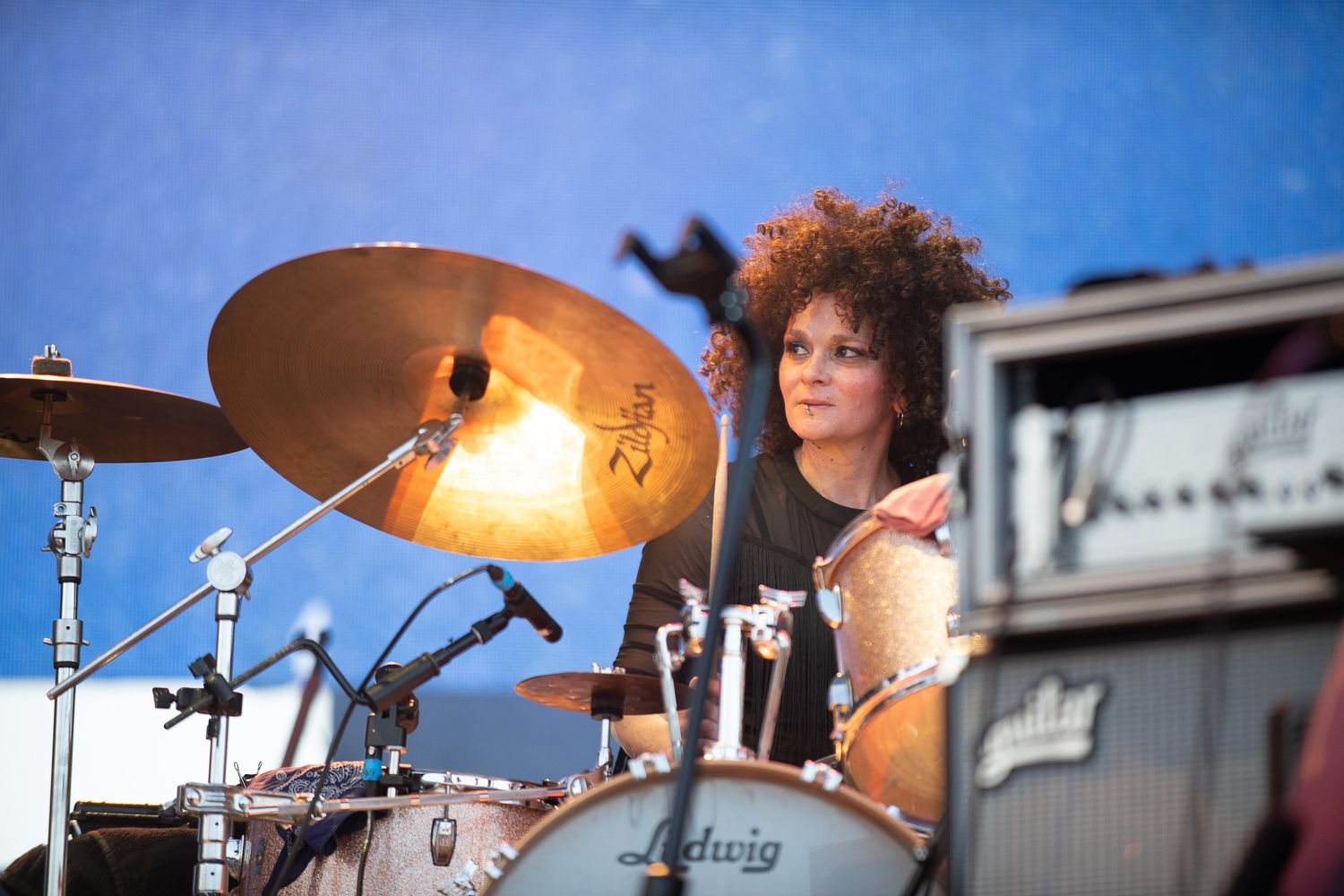 Drummer performs energetically behind a drum kit on stage at an outdoor music festival during a sunny day.