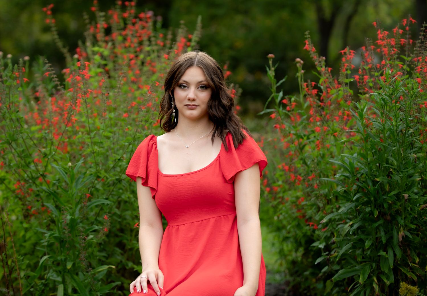A person in a red dress poses outdoors among coral-colored flowers in a garden setting.