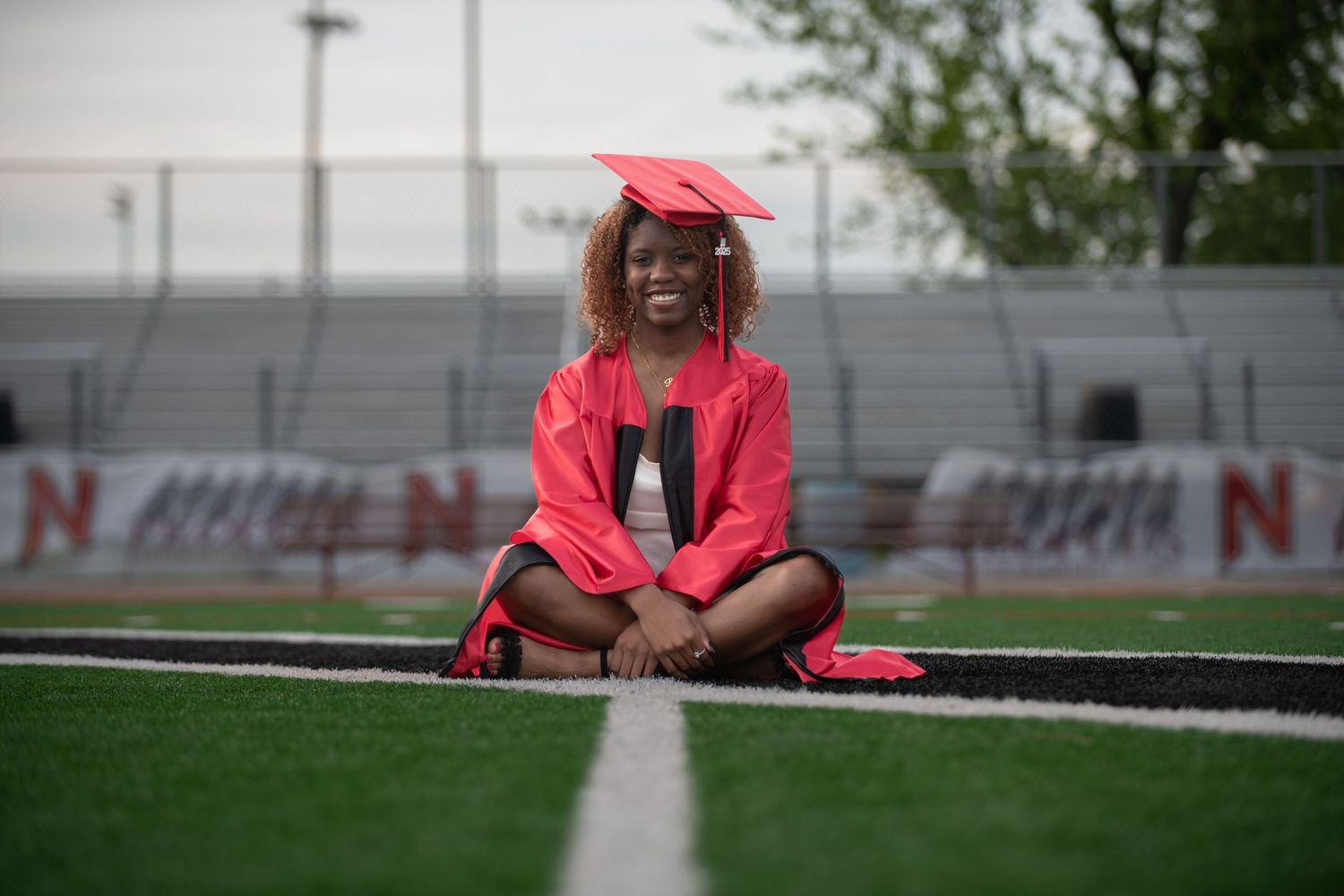 Figure in bright red graduation cap and gown sits on outdoor bleachers.