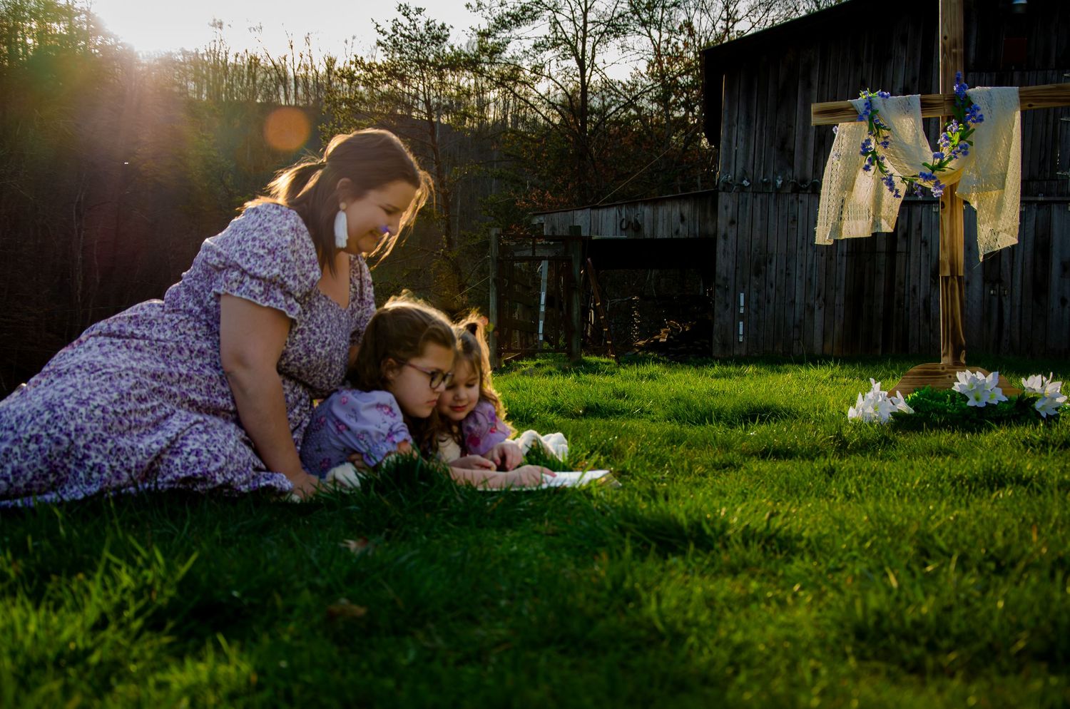 Mother and child relaxing on lush grass near a weathered barn during sunset.