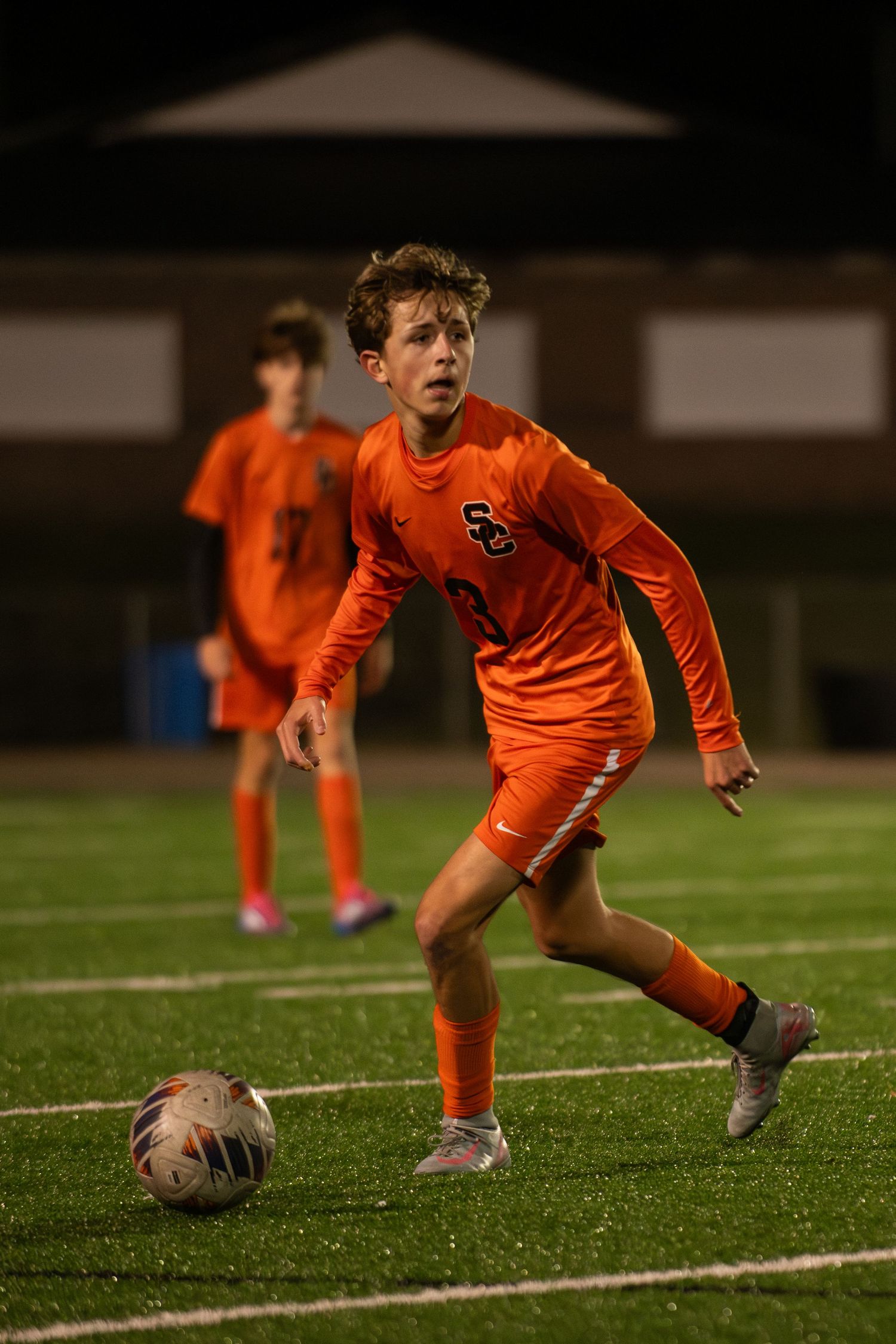 Young soccer player in orange uniform slides to control the ball during a nighttime match on artificial turf.