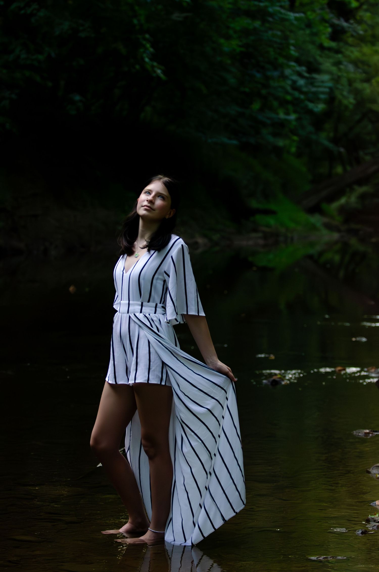 Woman in striped dress stands in shallow water during moody forest photoshoot.