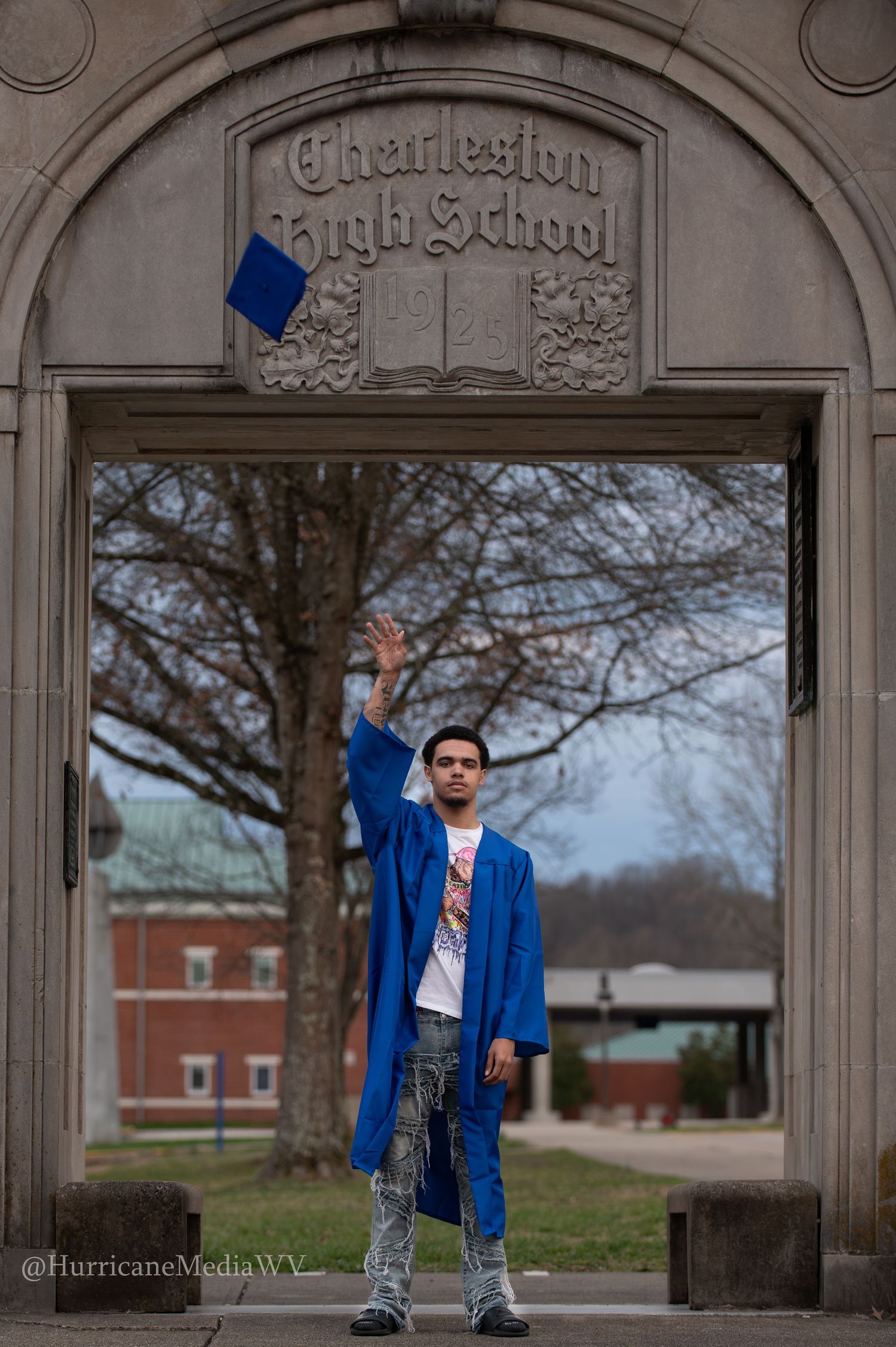 Graduate in blue cap and gown stands under historic Charleston High School stone archway during graduation photo shoot.