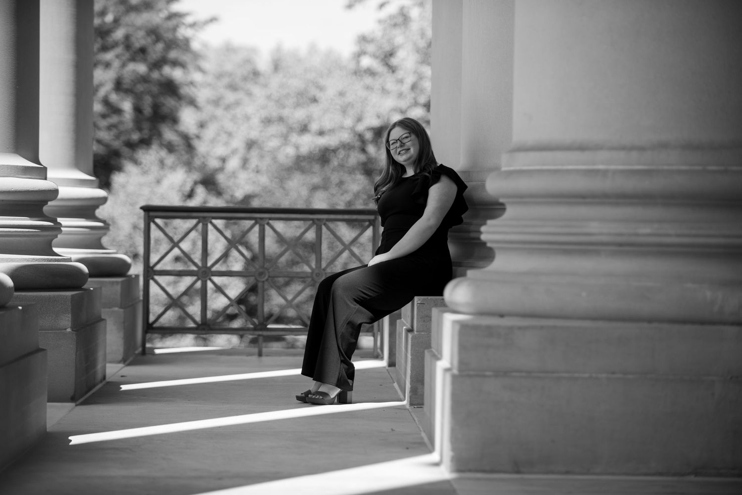 Artistic black and white photo of person sitting on architectural columns in dramatic lighting.