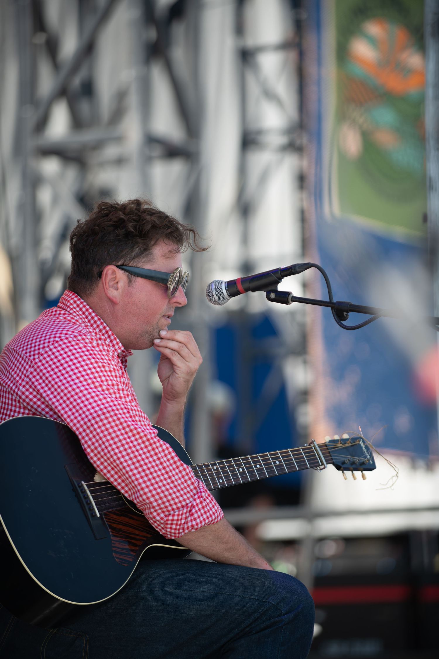 Musician in red checkered shirt plays acoustic guitar on outdoor stage in sequence of performance shots.