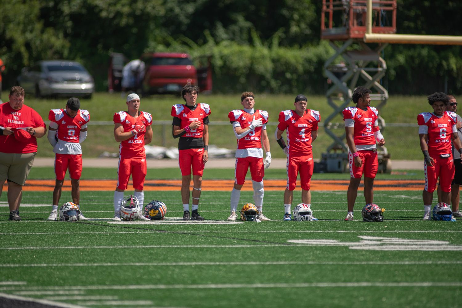 Youth football players in red uniforms line up on a green turf field during a sunny outdoor game day.