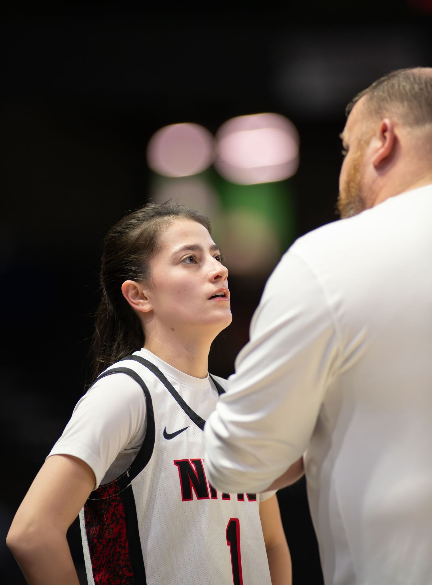A basketball player in a white Nike jersey looks up at their coach during a game conversation.