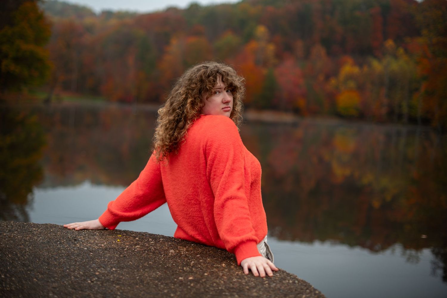 A person in a bright coral sweater poses near a serene autumn lake surrounded by colorful fall foliage.