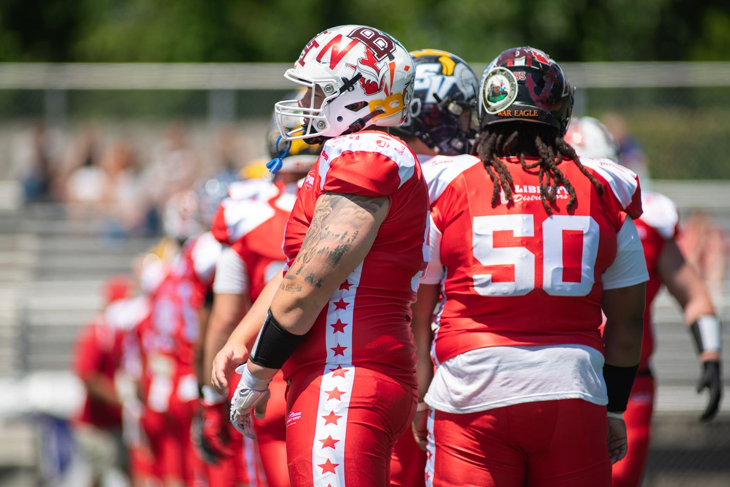 Football players in red All-Star uniforms stand on field, player #50 visible during warm-ups on sunny day.