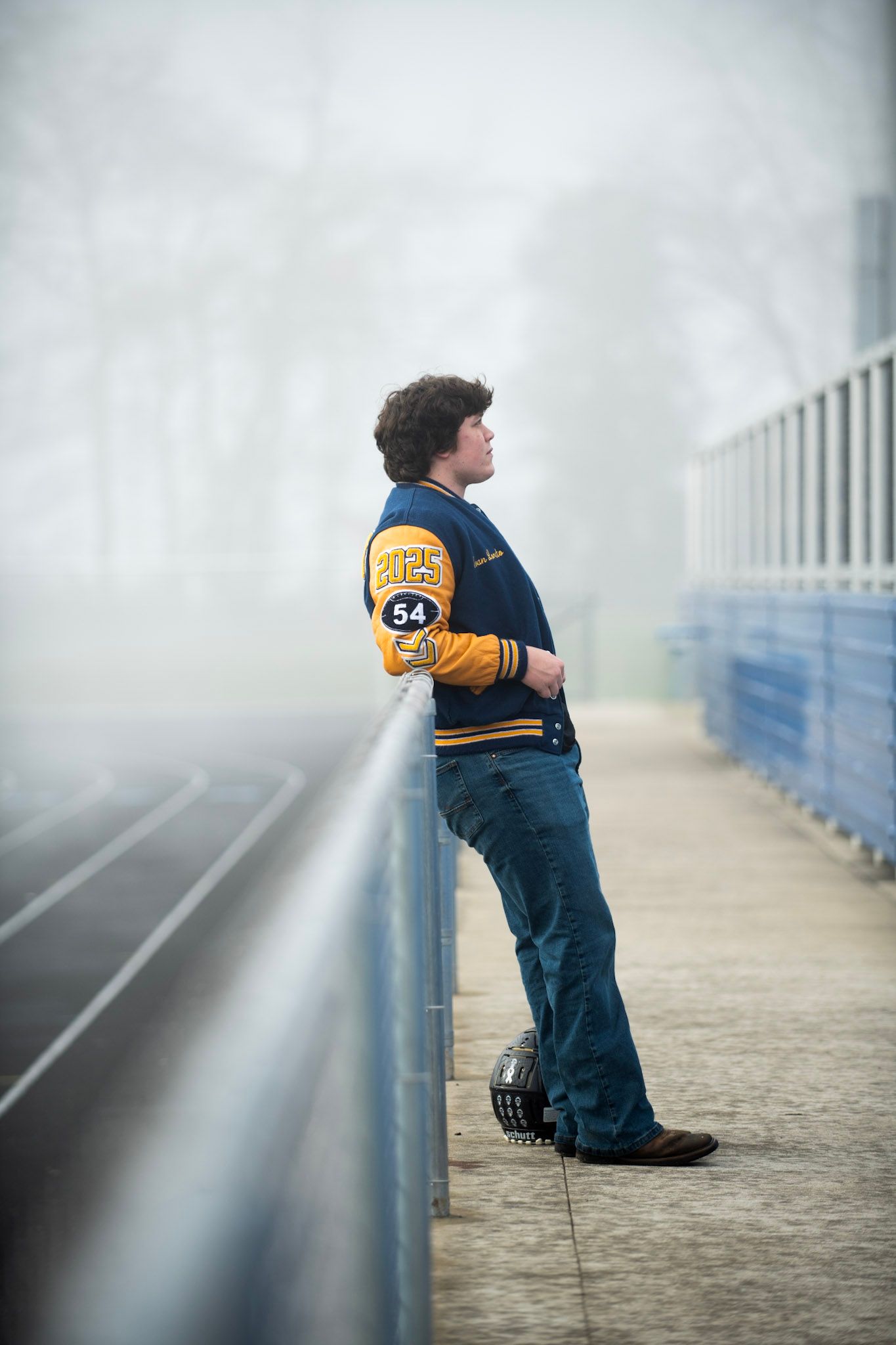 Someone in a varsity jacket leans against a foggy railing overlooking a track.