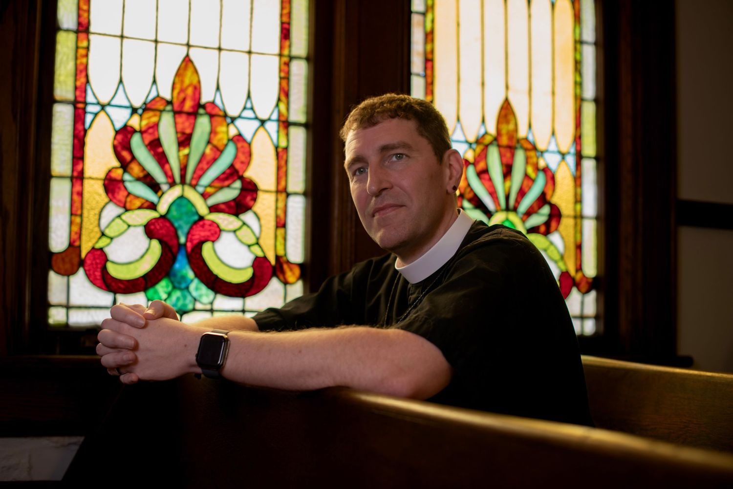 Person in black attire sitting in church pew with stained glass windows in background.
