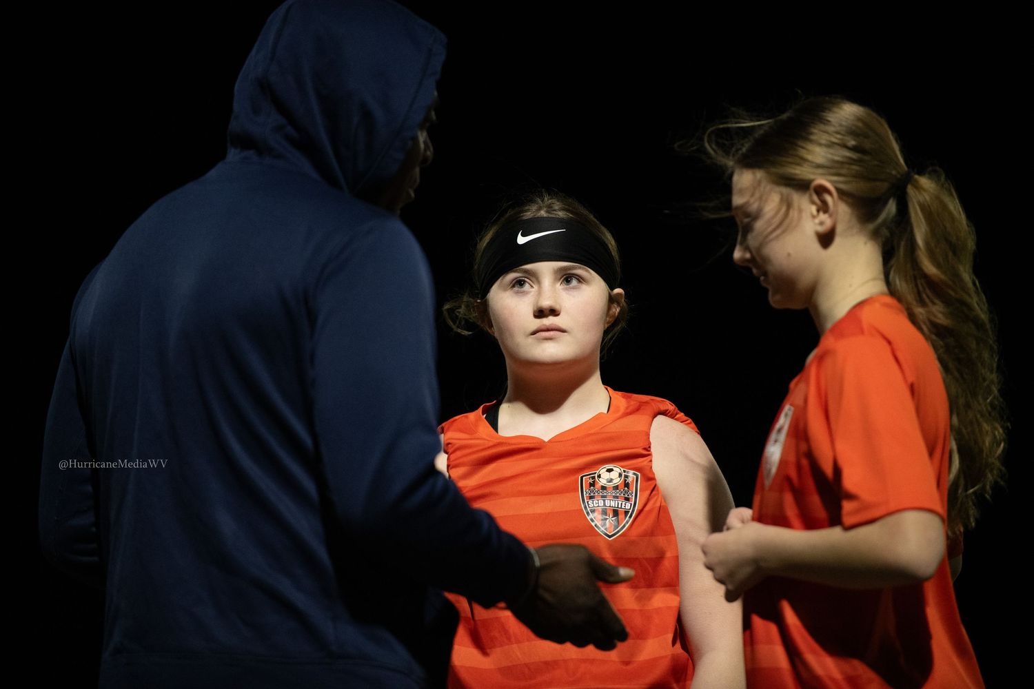 Athletes in orange jerseys huddle with coach during nighttime sports event.