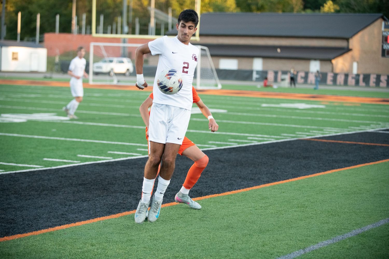 Soccer player in white uniform dribbles ball down sideline of artificial turf field during daytime match.