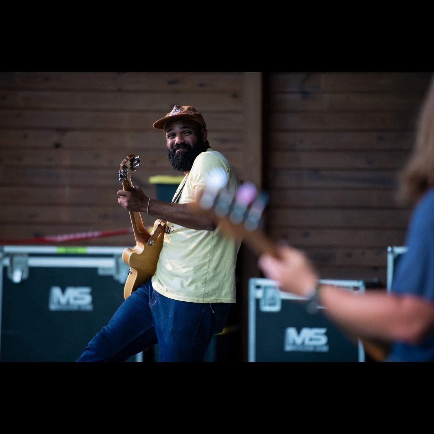 A person in a yellow shirt plays guitar on a dimly lit stage with performance equipment visible in the background.