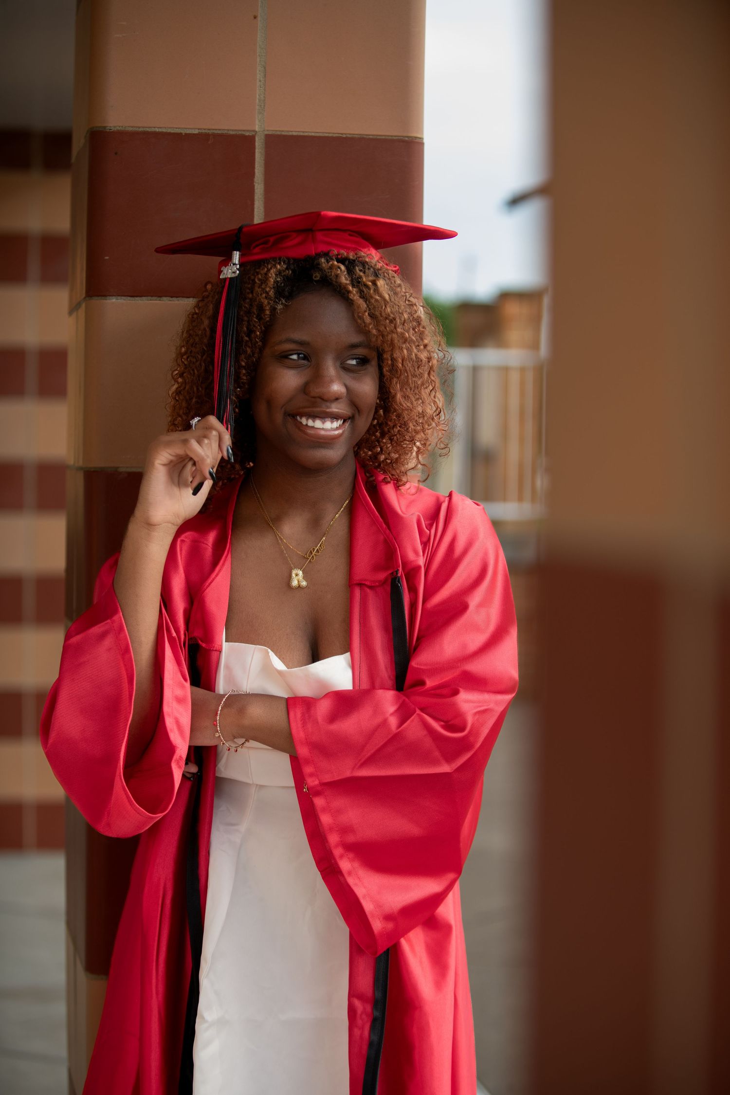 Graduate in red cap and gown smiles brightly while posing for graduation photos in academic hallway.