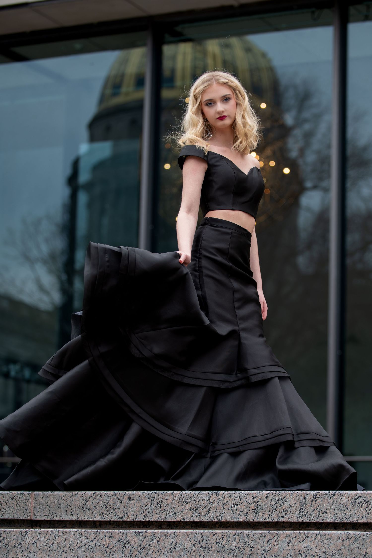 Woman in flowing black formal dress posing dramatically against modern building exterior.