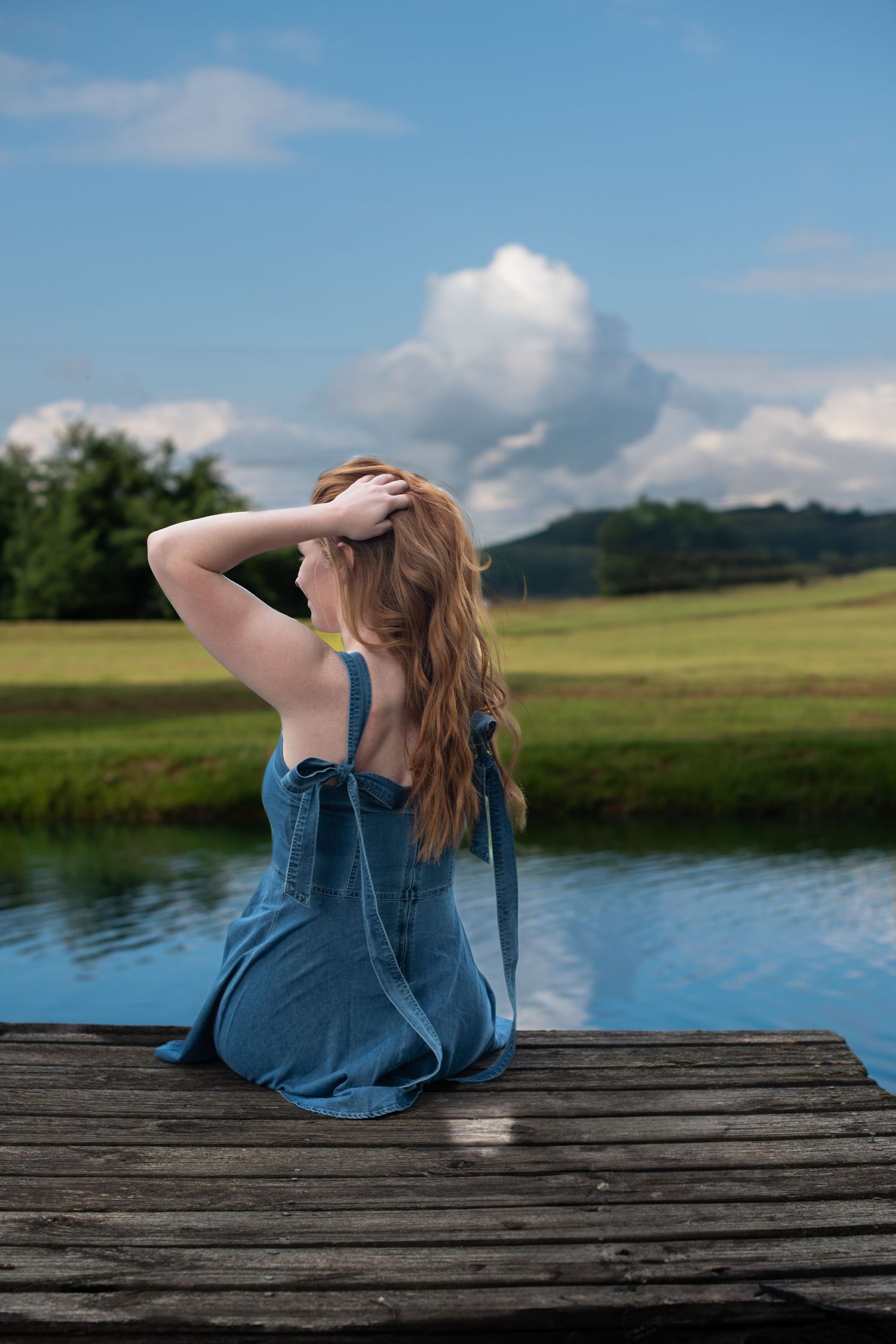 A person in denim sits on a wooden dock by a serene lake with rolling hills and blue sky in the background.