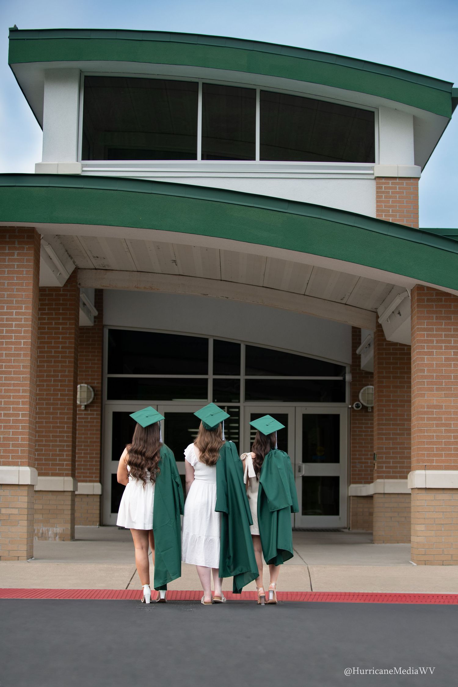 Students in green graduation gowns gather outside a modern school building entrance.