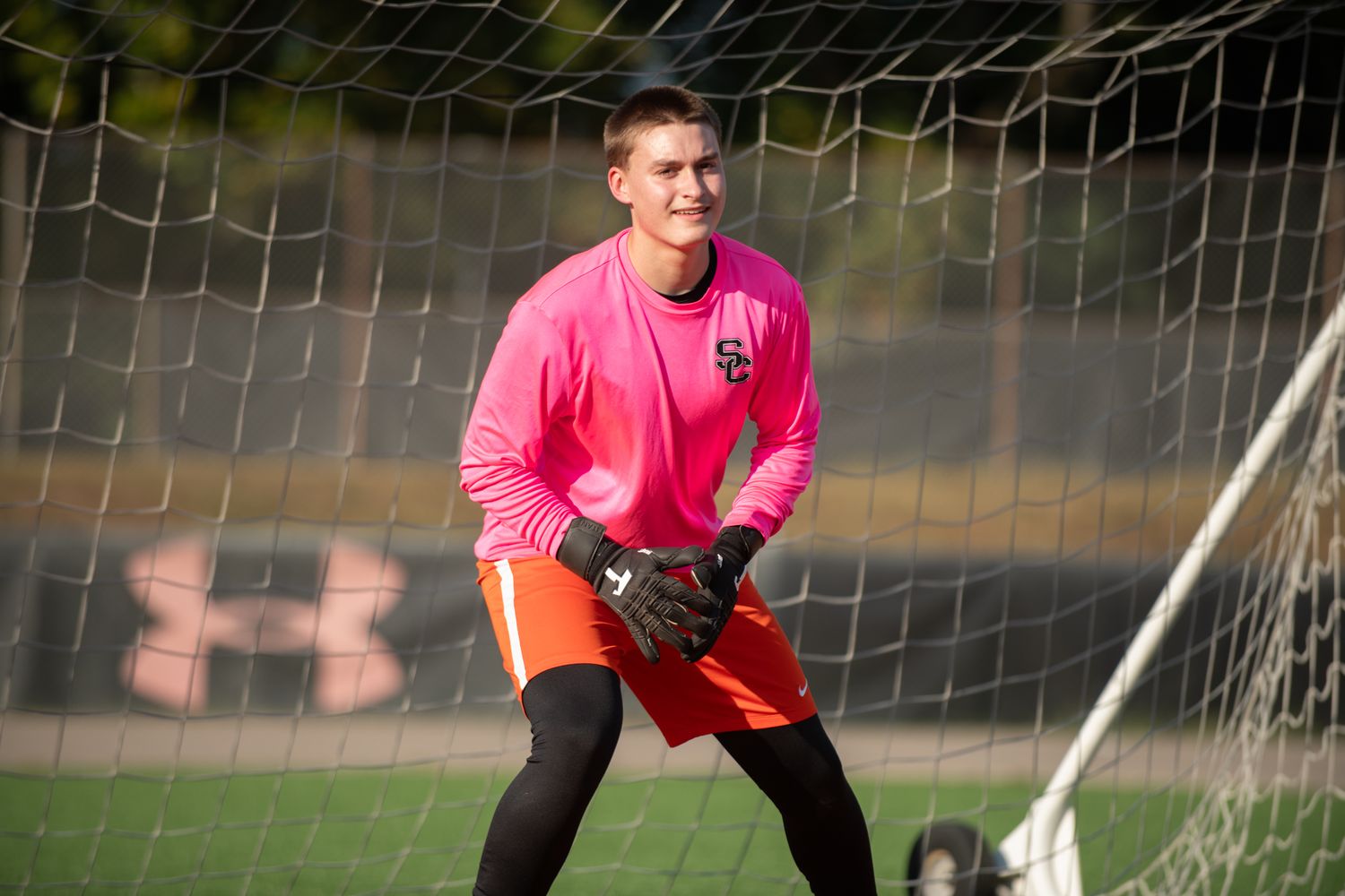 Soccer goalkeeper in pink jersey and orange shorts stands ready in defensive position in front of goal net.