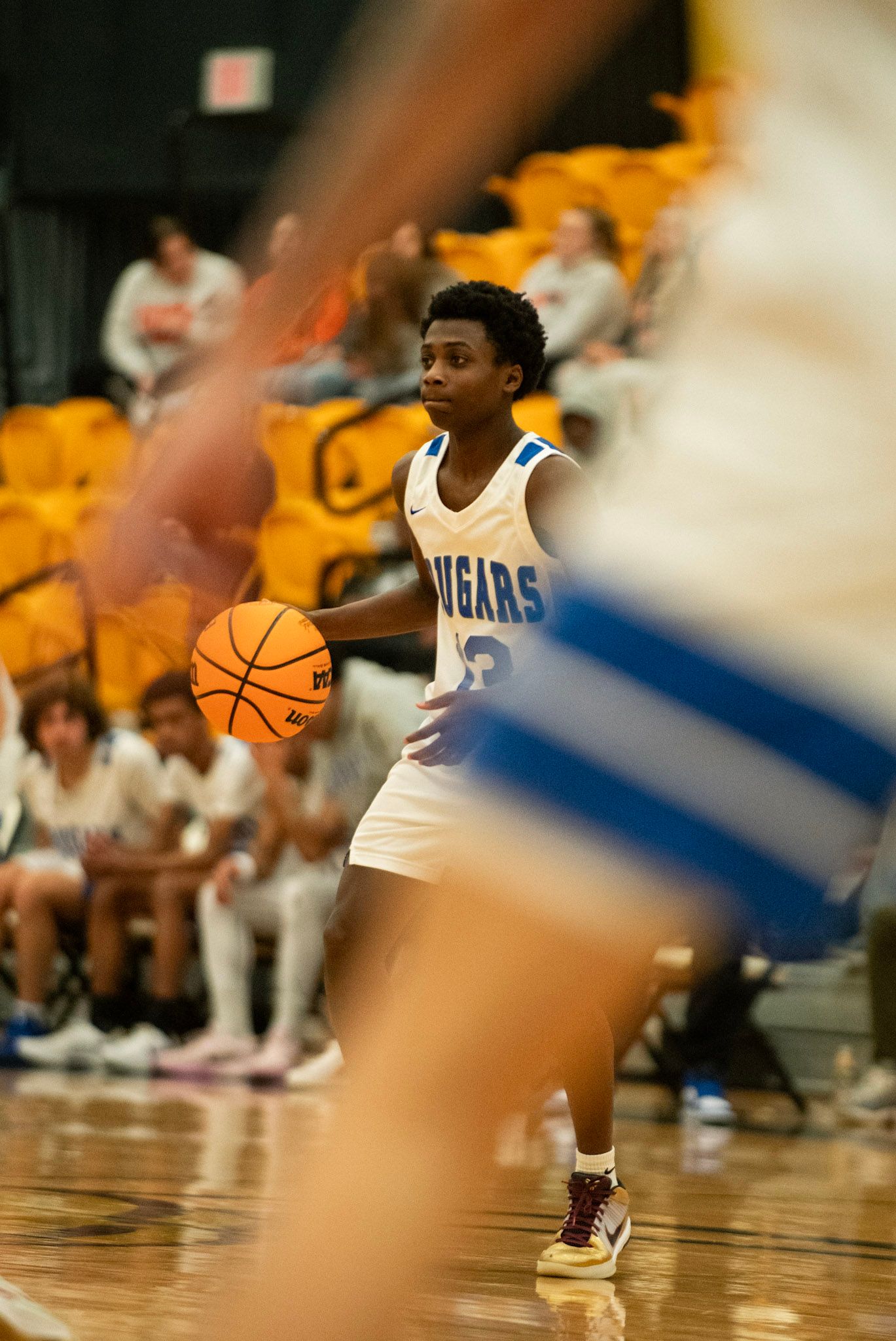 Basketball player in white Cougars jersey dribbles ball during game with yellow bleachers visible.