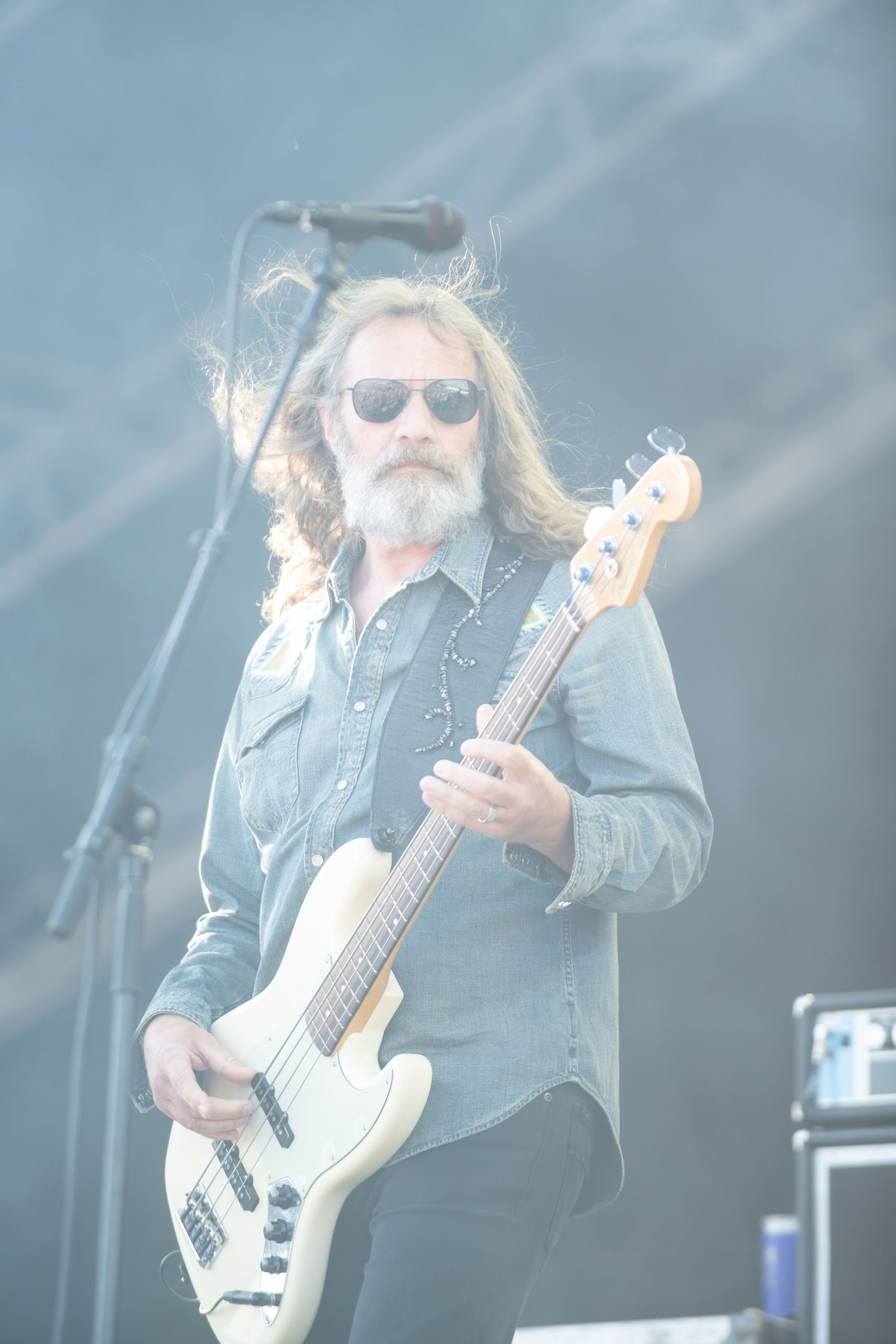 A musician in denim and sunglasses plays bass guitar on stage during a live performance in atmospheric lighting.